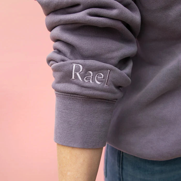 Close up of Rael embroidery on wrist of gray sweatshirt with the word 'Balanced' printed on the front, displayed on a pink background.
