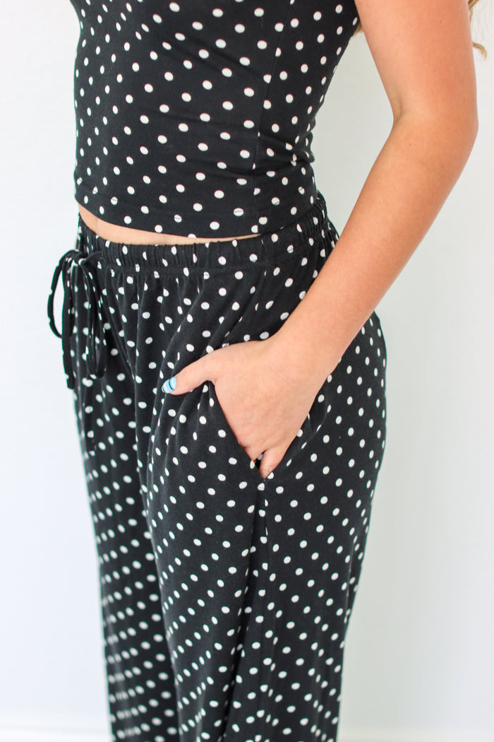 A person wearing a black two-piece set with white polka dots, featuring a sleeveless crop top and matching drawstring pants with side pockets.