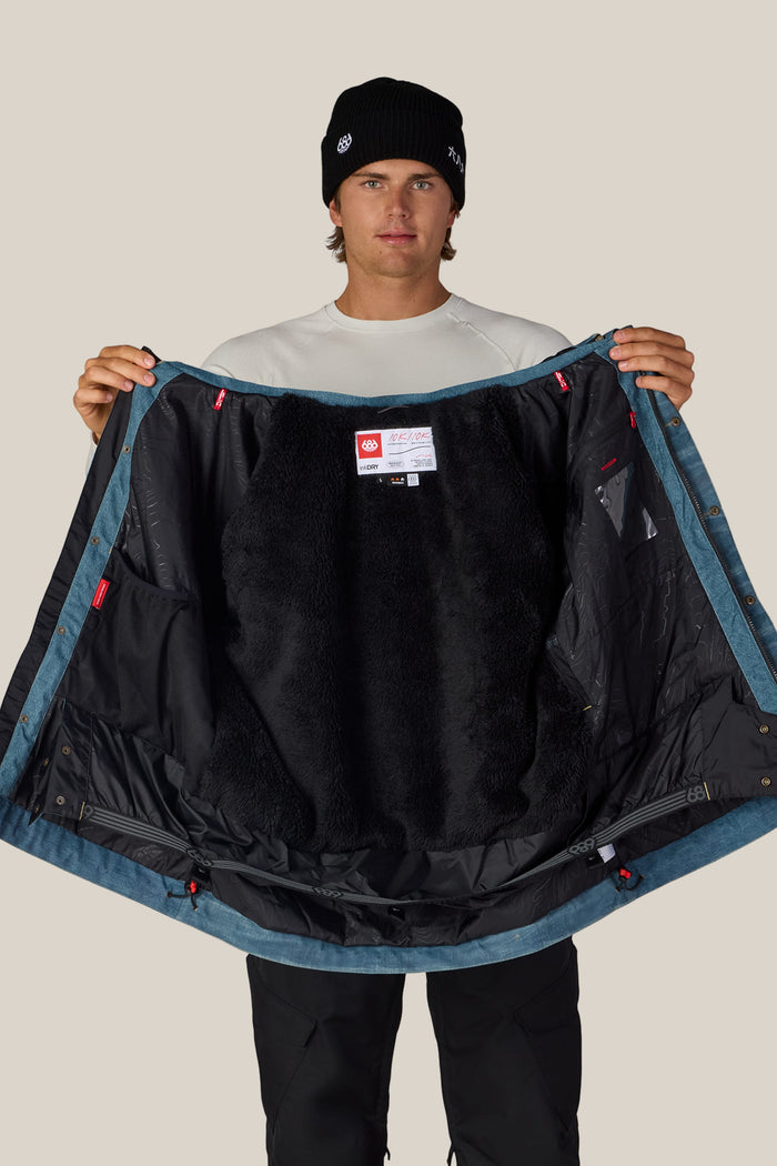 A person in a black beanie and white shirt holds open the 686 Men’s Deconstructed™ Denim Insulated Jacket, displaying its black furry lining and inner labels.