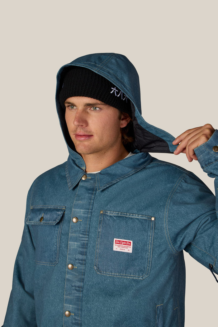 A person wearing the 686 Men’s Deconstructed™ Denim Insulated Jacket and a black beanie adjusts the jacket’s hood with one hand. The 686 denim jacket features metal buttons and a red and white label on the chest. The background is plain and light-colored.