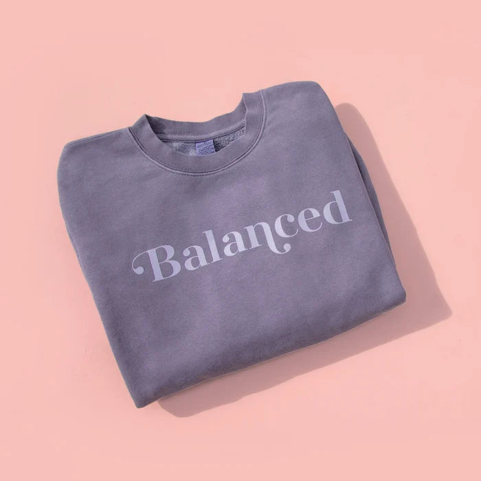 A gray sweatshirt with the word 'Balanced' printed on the front, displayed on a pink background.
