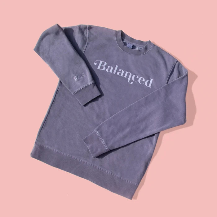 A gray sweatshirt with the word 'Balanced' printed on the front, displayed on a pink background.