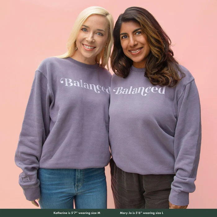 Two women wearing gray sweatshirt with the word 'Balanced' printed on the front, displayed on a pink background.