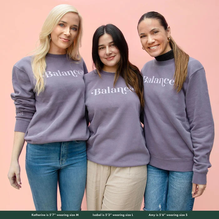 Two women wearing gray sweatshirt with the word 'Balanced' printed on the front, displayed on a pink background.