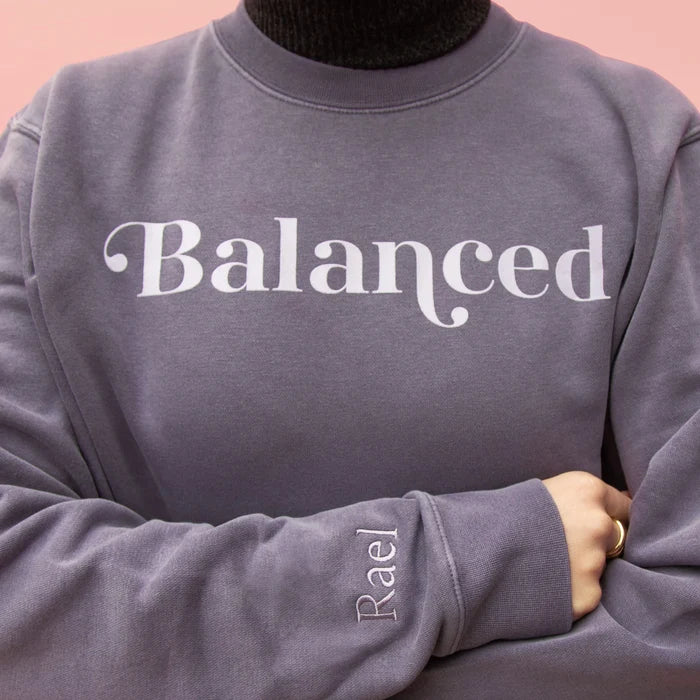 Close up of chest graphic and Rael embroidery on wrist of gray sweatshirt with the word 'Balanced' printed on the front, displayed on a pink background.