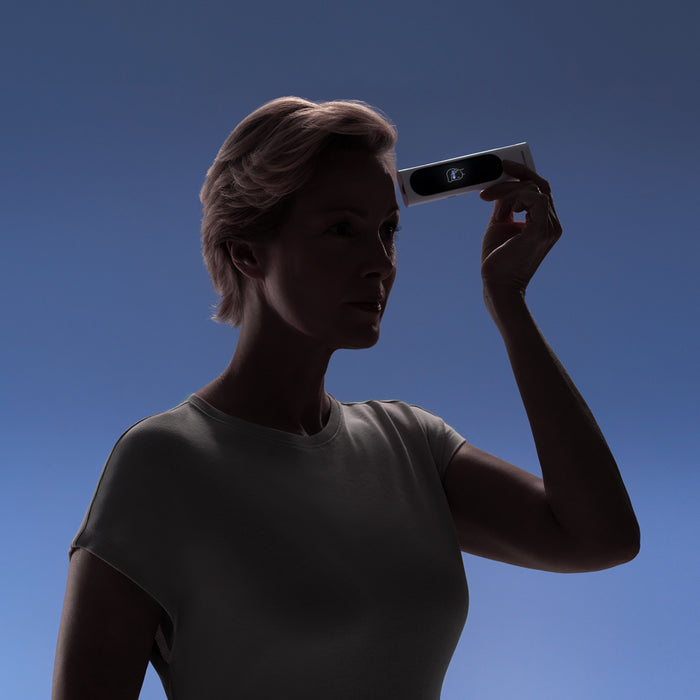 BeamO — Beam-O — Woman holding the device against her forehead, dramatic side lighting against blue gradient background, mid-body framing — casual outfit, forehead scan, shadow