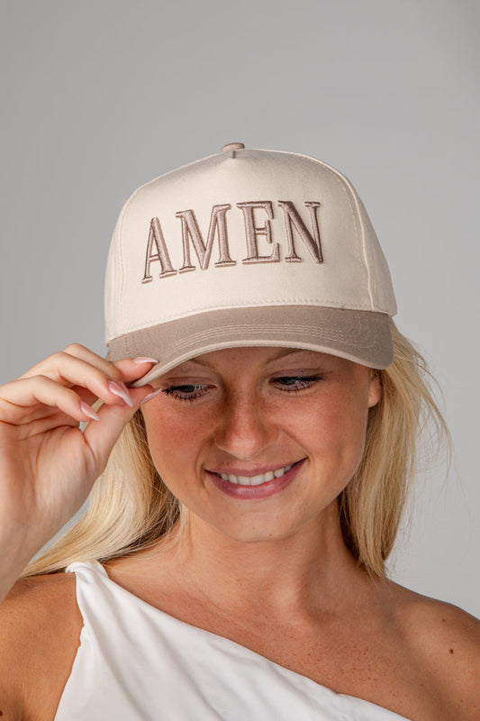 A woman wearing a beige baseball cap with 'AMEN' embroidered in brown across the front, photographed against a plain light gray studio background.