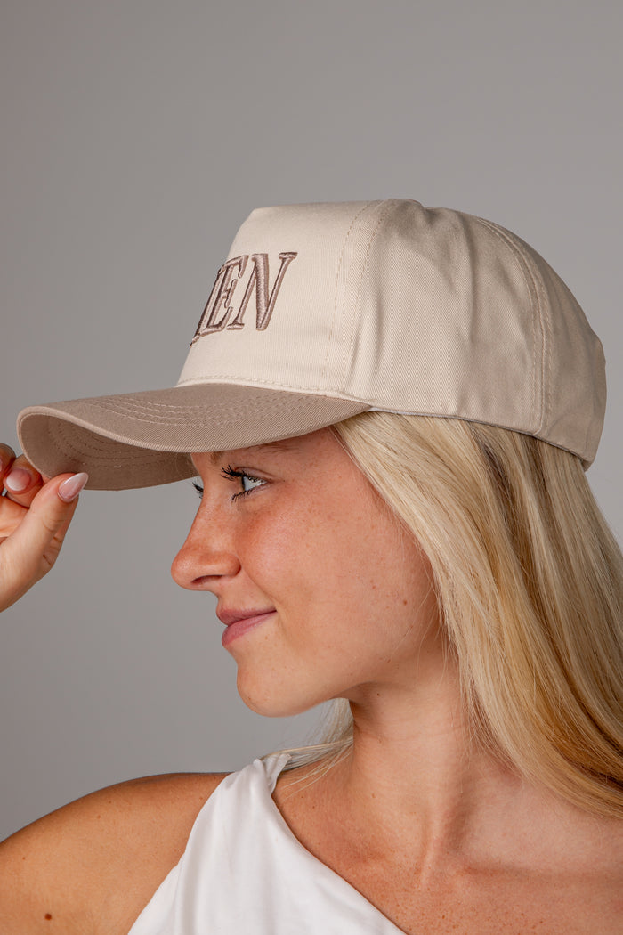 A woman in profile wearing a beige baseball cap with 'AMEN' embroidered in brown on the front, photographed against a plain gray studio background.