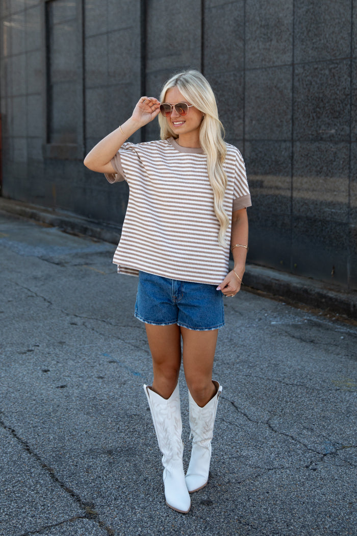 A woman stands outdoors wearing a beige and white horizontally striped short-sleeve t-shirt, blue denim shorts, and tall white cowboy boots, posing in front of a dark gray stone wall.