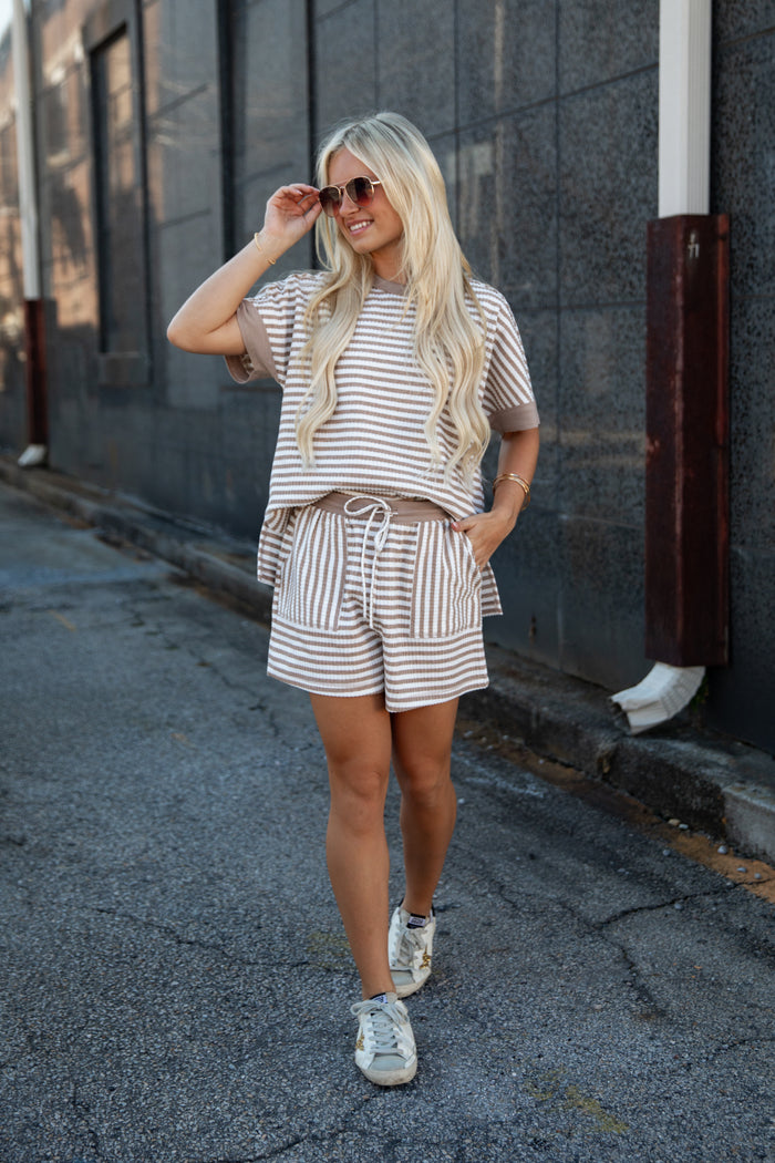 A woman wearing a matching beige and white striped lounge set with a short-sleeve top and shorts, walking down an outdoor alleyway.