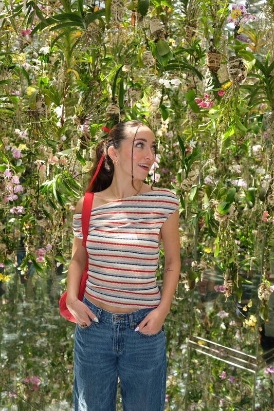 Woman standing in a garden with flowers and plants around her wearing a stripe off the shoulder top