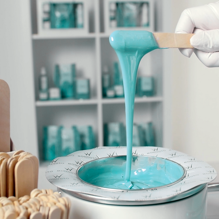 W383 Italwax 1kg Vanira Spa Sandalwood Film Wax | Aqua Blue Italwax Vanira Sandalwood Film Wax being poured from a wooden spatula into a wax heater with shelves of similar products in the background.