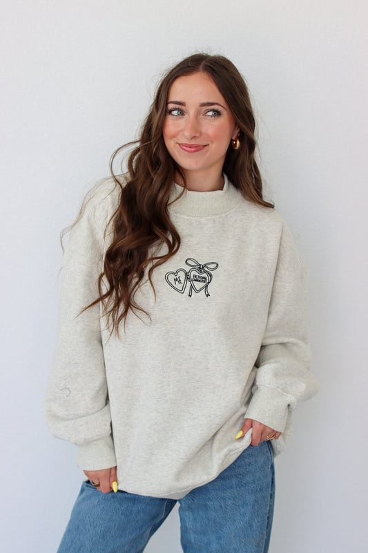 Woman wearing a light gray crewneck with a embroidery design against a white background