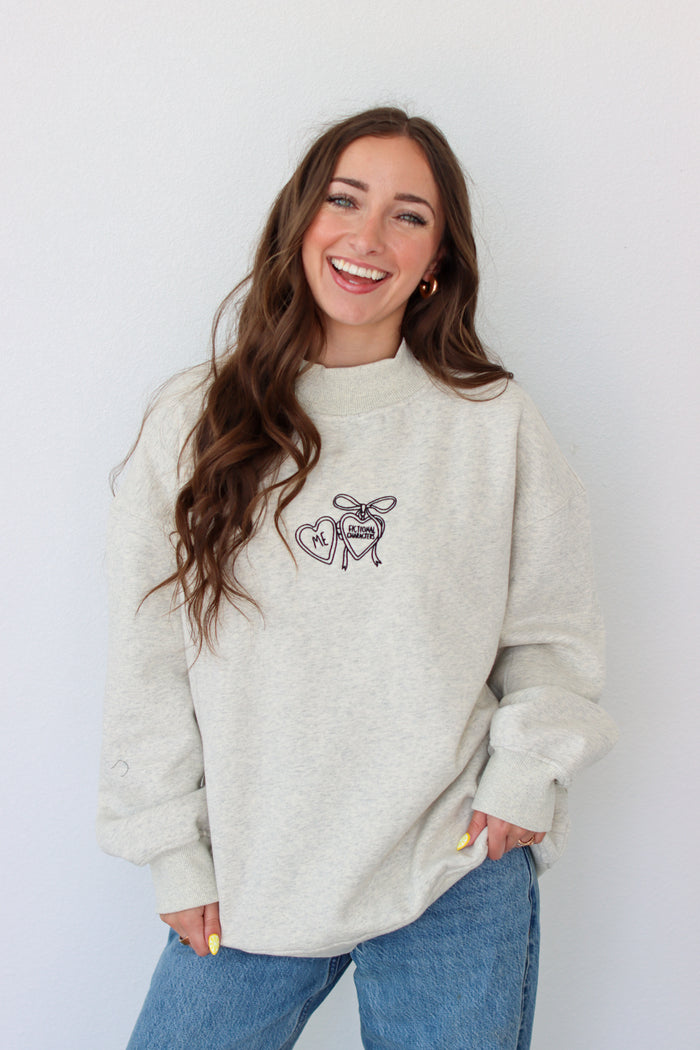 Woman wearing a light gray crewneck with a embroidery design against a white background