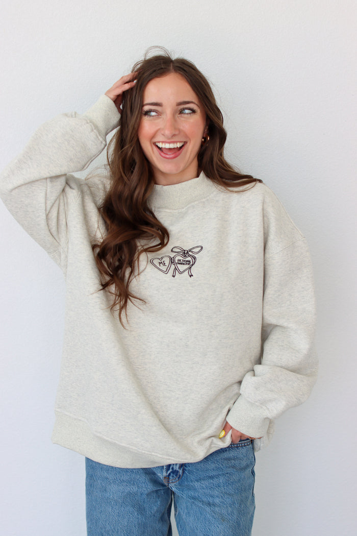 Woman wearing a light gray crewneck with a embroidery design against a white background