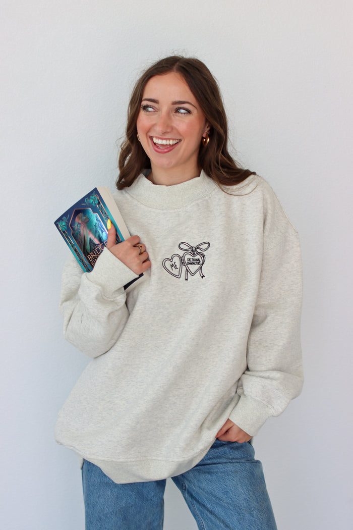 Woman wearing a light gray crewneck with a embroidery design against a white background