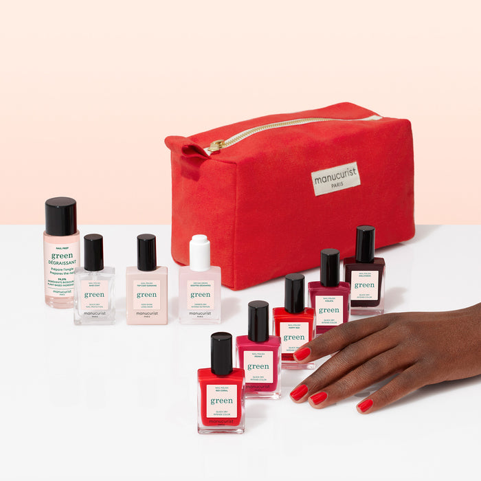 The Manucurist Super Starter Kit, including several plant-based nail polishes and treatments, is shown on a white surface with a red makeup bag. A hand with red gel-effect nails rests beside the products.