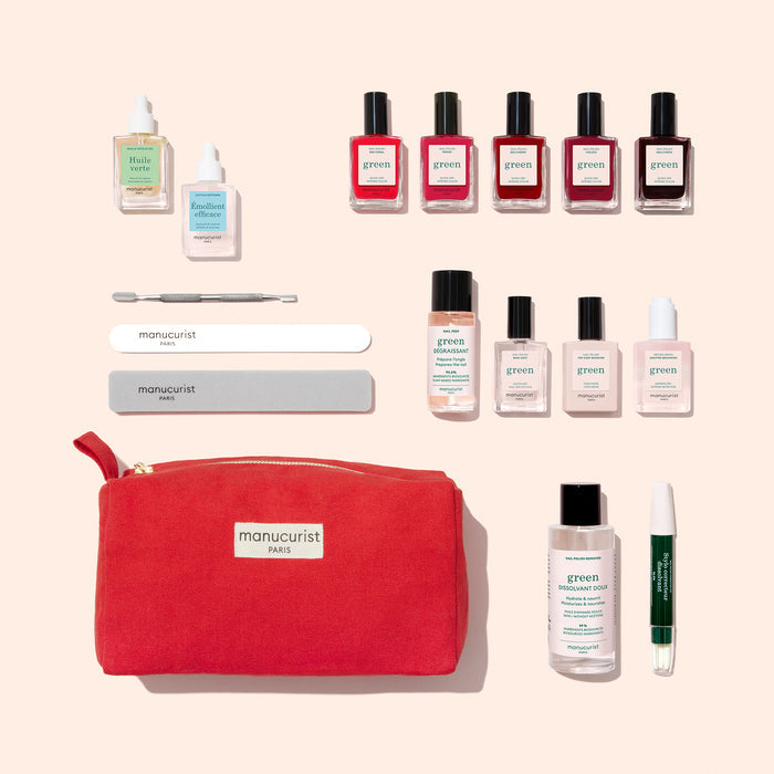 The Manucurist Super Ultimate Kit is shown in a flat lay with a red pouch, colorful nail polishes, nail prep kit, care bottles, cuticle pusher, nail files, green pen, and small brush—everything for an at-home manicure on a beige background.