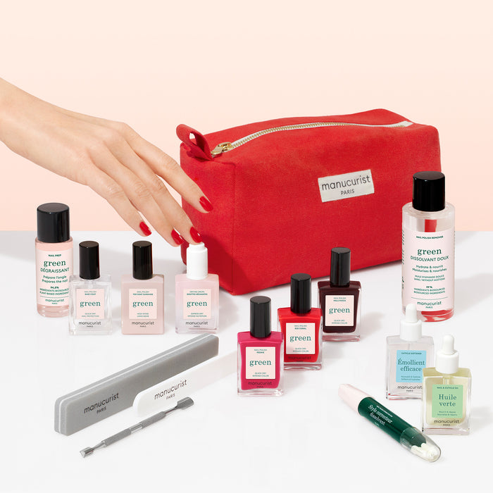 A hand with red nails reaches for the Manucurist Ultimate Kit, featuring a red zippered bag surrounded by gel effect polishes, files, glass bottles, and other nail care essentials on a white surface.